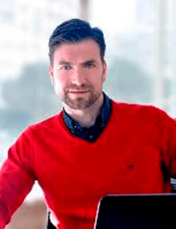 A man in a red sweater and blue shirt standing in an office environment with a laptop in front of him.