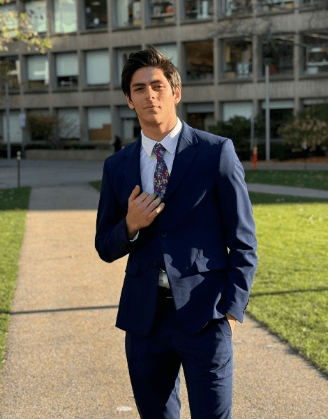 A young man in a blue suit adjusting his tie, standing outdoors with buildings in the background.