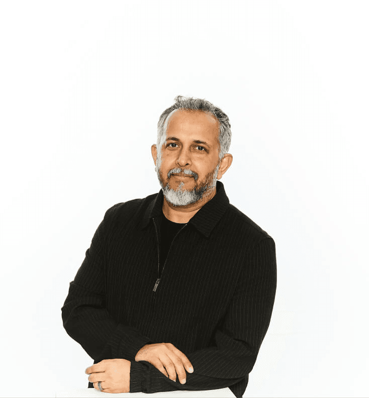 A middle-aged man with gray hair and a beard stands confidently with his arms crossed, wearing a black zip-up sweater, against a white background.