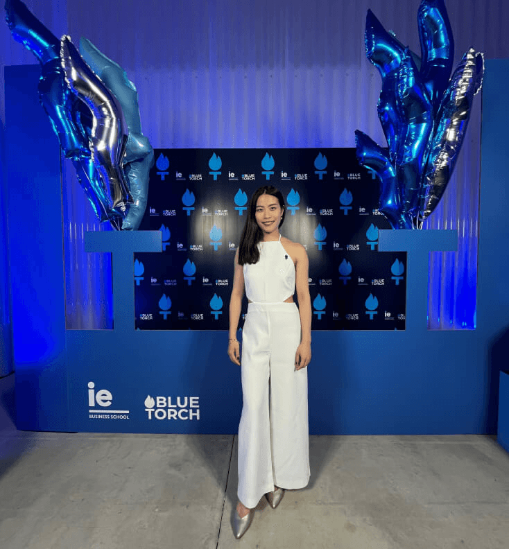 A woman in a white jumpsuit is standing in front of a promotional backdrop with blue balloons and logos.