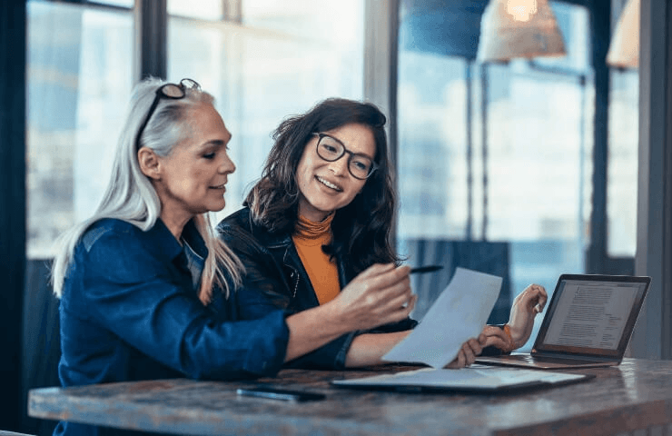 Two women are reviewing documents and working on a laptop in a modern office setting.