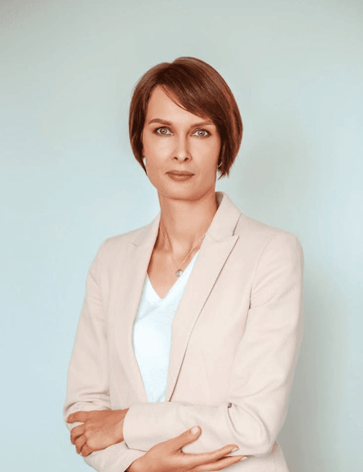 A professional looking woman with short hair stands confidently with her arms crossed, wearing a light pink blazer over a white blouse against a pale blue background.