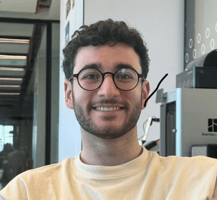 A young man with curly hair and glasses, smiling in an indoor environment.